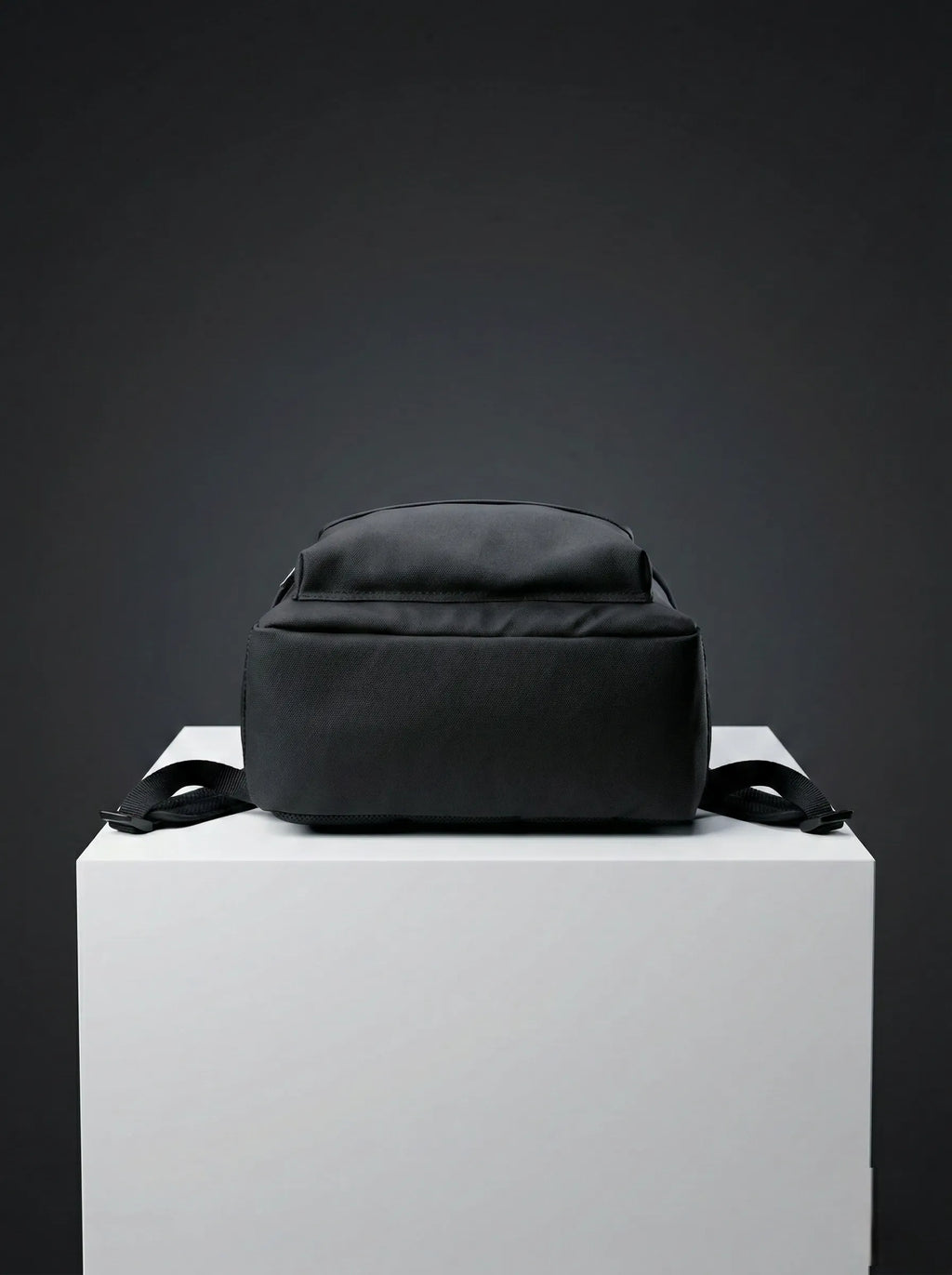 Bottom view of black EQ1 backpack placed on a white display platform with clean black fabric and adjustable side straps.