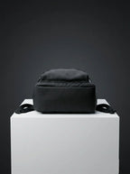 Bottom view of black EQ1 backpack placed on a white display platform with clean black fabric and adjustable side straps.