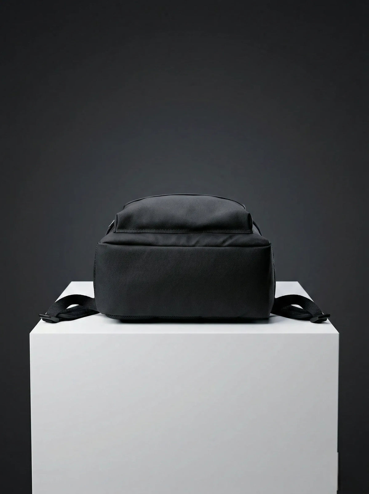 Bottom view of black EQ1 backpack placed on a white display platform with clean black fabric and adjustable side straps.